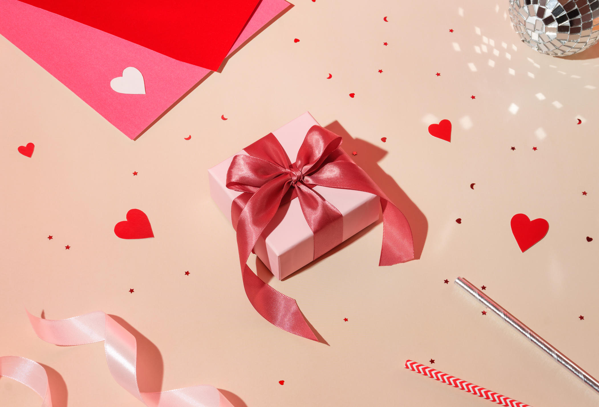 Valentine day composition concept: white gift box with magenta colored bow, red hearts confetti and pink paper on pastel background. High angle view, flat lay 