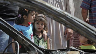Zahra Naaim, 4, center, and Balqes Youssif, 6, left, stand next to a destroyed vehicle in the Shiite stronghold of Sadr City in Baghdad, Iraq, Saturday, April 24, 2010. A series of bombings mainly targeting Shiite worshippers killed at least 69 people on  