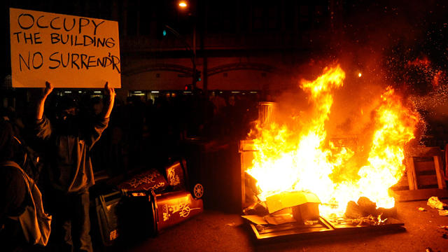Occupy Oakland protester raises a sign near a burning trash heap  