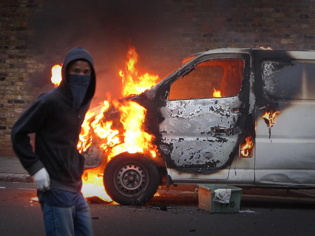 A hooded youth walks past a burning vehicle 