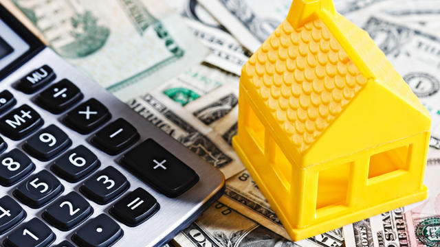 A yellow toy house sits on a background of many US dollars, next to a calculator. 