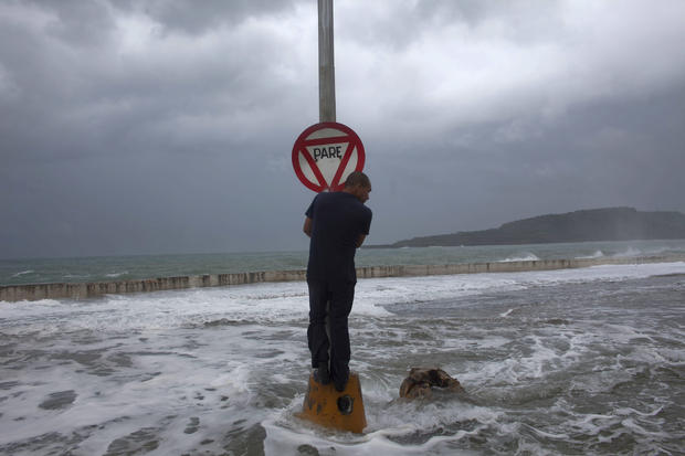 A man stands on a post with a stop sign as waves pass the seawall 