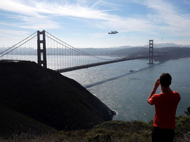 A spectator takes a photo of the Space Shuttle Endavour as it flies on top of a modified 747 jumbo jet over the Golden Gate Bridge while traveling to Los Angeles on September 21, 2012 in Sausalito, California. The Space Shuttle Endeavour did a 4-1/2 hour tour over California landmarks before heading to Los Angeles International Airport where it will be prepared to be moved to its new permanent home at the California Science Center in downtown Los Angeles. The shuttle will be on public display starting October 30. (Photo by Justin Sullivan/Getty Images) 
