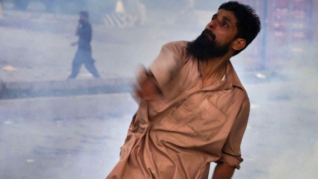A Pakistani protester hurls back a tear gas canister fired by police, as another protester talks on a cell phone in the background 