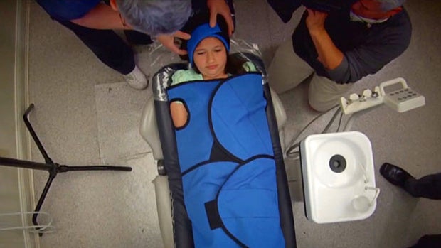 ​Dr. Mark Burhenne, left, demonstrates how a papoose board immobilizes a patient for a segment broadcast on CBS Boston station WBZ-TV on May 17, 2016. 