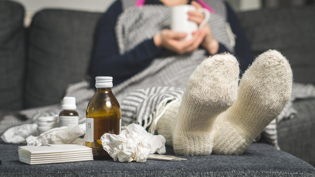 Cold medicine and sick woman drinking hot beverage to get well from flu, fever and virus. Dirty paper towels and tissues on table. Ill person wearing warm woolen stocking socks in winter. 