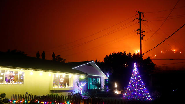 People stand on a roof of a home illuminated with Christmas lights to watch wildfire on a hillside burn during the Thomas Fire in Santa Barbara county near Carpinteria 