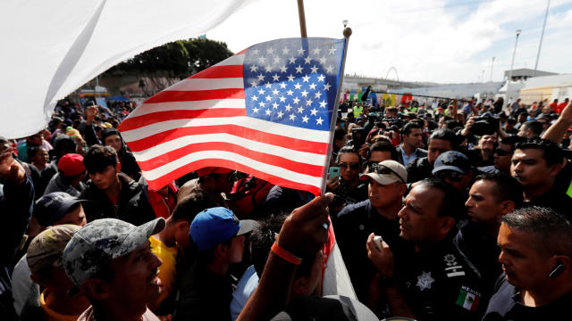 A migrant, part of a caravan of thousands traveling from Central America to the United States, holds a U.S. flag as the caravan negotiates with Mexican policemen near the El Chaparral port of entry of border crossing between Mexico and the United States i 