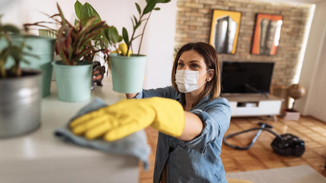 Woman wiping dust from shelf and other furniture in living room 