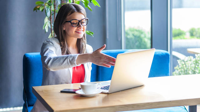 Portrait of happy beautiful stylish young woman in glasses sitting, looking at her laptop screen on video call and giving hand to handshake, toothy smile. 