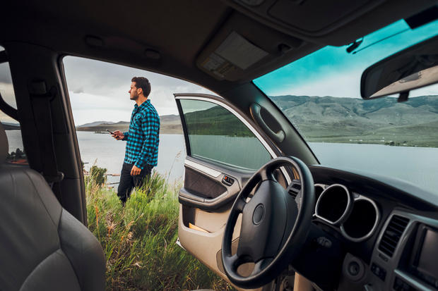 Mid adult man standing beside Dillon Reservoir, holding smartphone, view through parked car, Silverthorne, Colorado, USA 