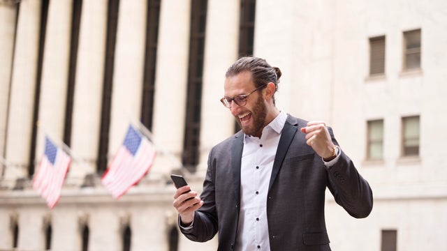 Excited young businessman looking at cell phone in front of Stock Exchange, New York City, USA 