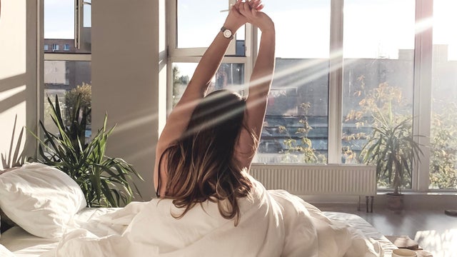 Rear View Of Young Woman With Arms Raised Sitting On Bed At Home 