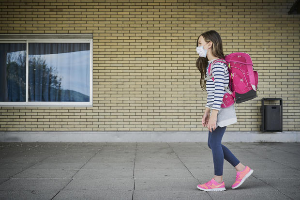 Girl wearing face mask and schoolbag walking along building 