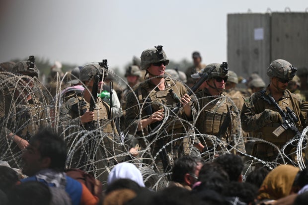 U.S. soldiers stand guard behind barbed wire as Afghans sit on a roadside near the military part of the airport in Kabul on Aug. 20, 2021.