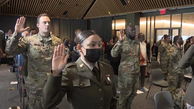 A room full of people, many in military uniforms, hold up their right hands during a naturalization ceremony 
