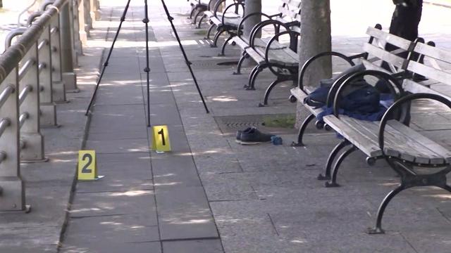 Two police markets sit on the ground in front of a bench in a park. There is a pair of shoes sitting next to the bench and what appears to be a blanket on the bench. 