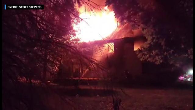 Flames shoot through the roof of a home surrounded by trees as a firefighter tries to spray the fire with a hose. 