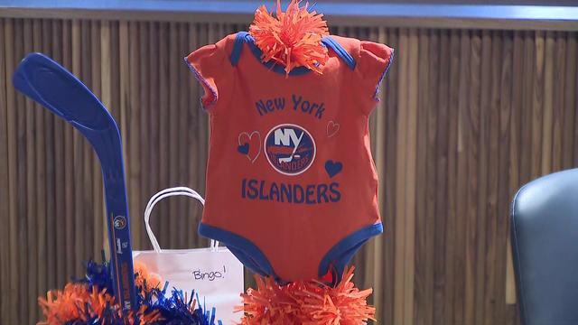 An orange baby onesie with blue trim, blue hearts and the New York Islanders logo is on display on a table, along with blue and orange pom-poms and a blue hockey stick. 