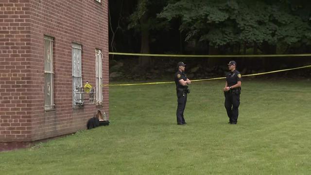 Two police officers stand on a lawn next to an apartment building. There is caution tape cordoning off a section of the yard. 