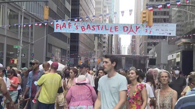 A large crowd of people fills the street beneath a banner reading "FIAF's Bastille Day 2022" and a string of small French flags. 