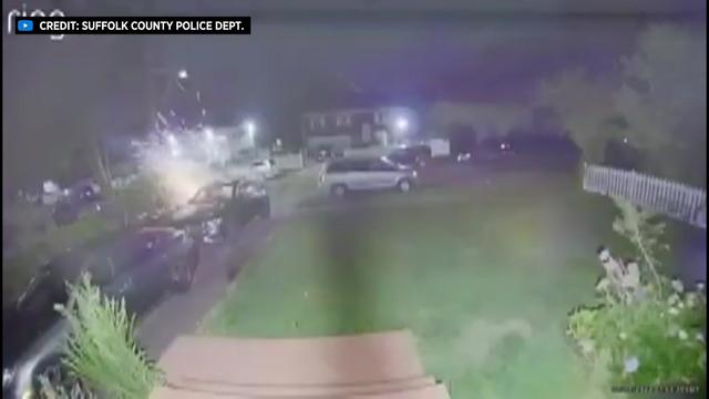 Video taken from the front door of a home shows two cars parked in a driveway and several cars parked on the street in front of the house. A large explosion is seen on the side of one of the cars parked in the driveway. 