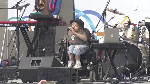 An individual in a wheelchair speaks into a microphone on a stage in front of a keyboard and a drum kit. 