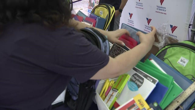 A volunteer stands in front of a table full of school supplies with an empty backpack. 