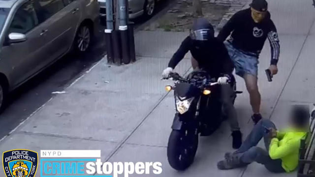 A still from surveillance video shows two individuals on a motorcycle. The passenger is pointing a firearm at an individual sitting on the sidewalk. 
