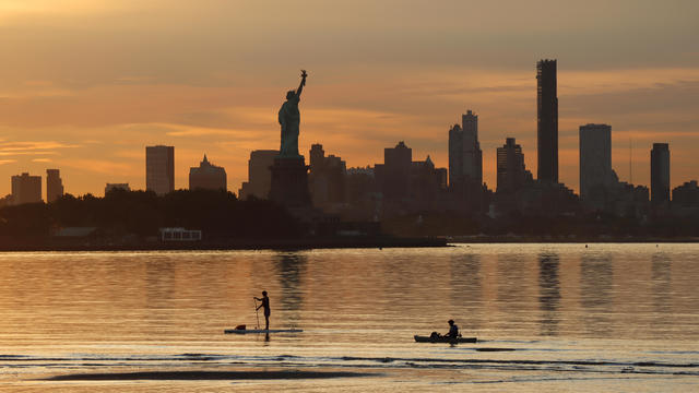 Two people paddle past the Statue of Liberty and the skyline of Brooklyn as the sun rises in New York City on August 15, 2022, as seen from Jersey City, New Jersey. 
