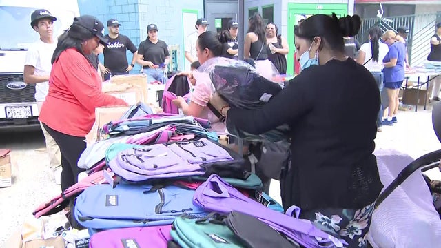 Adults and children stand at a table full of backpacks. 