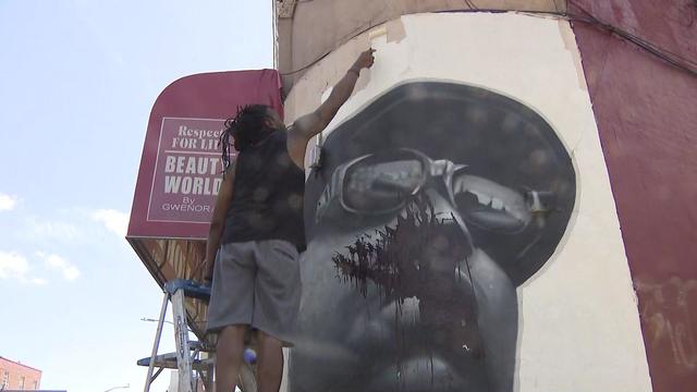 A man on a later paints a mural of Biggie Smalls on the outside corner of a building. Part of the mural has been splattered with dark-colored paint. 