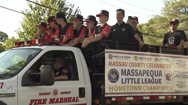 Players and staff from the Massapequa Coast Little League team stand in the back of a truck traveling along a parade route. 