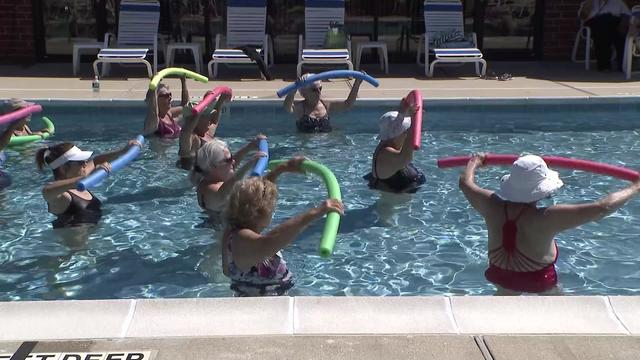 A group of seniors with pool noodles do water aerobics in an outdoor, in-ground swimming pool. 