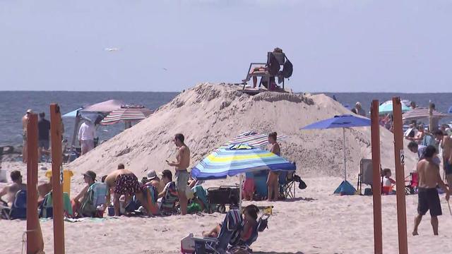 People sit in beach chairs in the sand next to a lifeguard seated atop a mound of sand. 