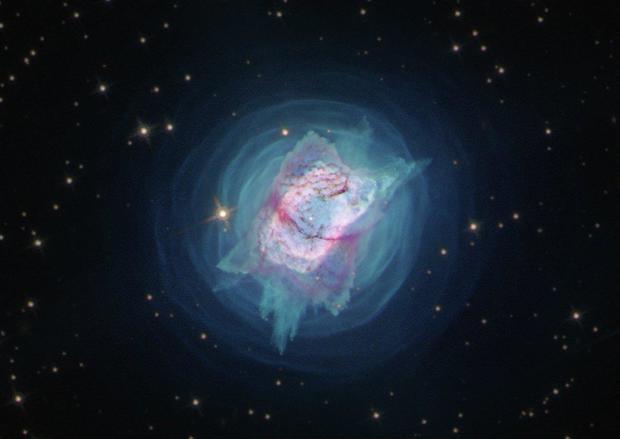 A nebula with a pink and blue pillow-shaped structure at a 45-degree angle in the middle, surrounded by round,  concentric waves of blue gas, all set on a black star-filled background. The pillow shaped structure has vein-like filaments of dark red gas and dust and a small bright star in the middle. 