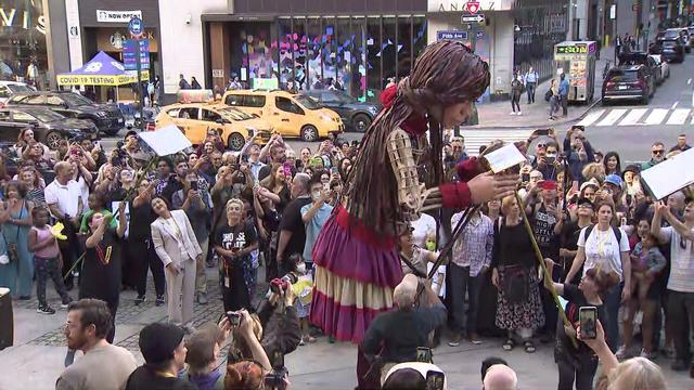 Dozens of people stand on a New York City sidewalk around a 12-foot tall puppet of a 10-year-old girl. Other puppeteers holding books on sticks surround her. 