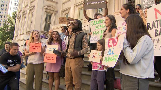 Adults and children stand outside the Department of Education headquarters, holding miscellaneous signs, some reading, "Let them play" and "End all mandate." 