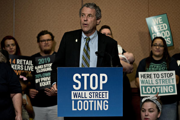 Sherrod Brown speaks at a podium surrounded by protesters 