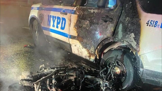 The charred remains of a moped lay on the ground next to an NYPD vehicle with several fire damage to its rear passenger door. 
