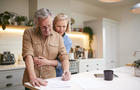 Mature Couple Reviewing And Signing Domestic Finances And Investment Paperwork In Kitchen At Home 