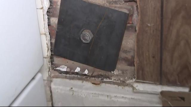 A hole in a wall covered by a black square piece of material and a large bolt. 