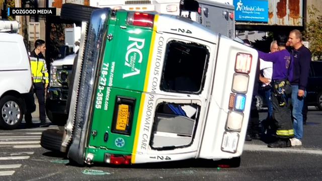 An ambulance with broken rear windows lies on its side in the street. 