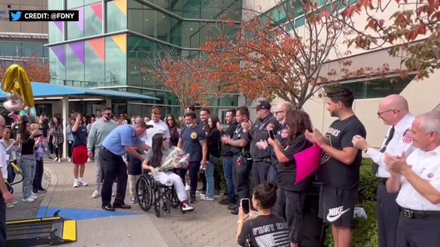 A woman in a wheelchair holds a bouquet of flowers while a crowd surrounds her, applauding. 