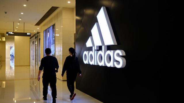 Two people walk past the big logo of Adidas in a shopping 