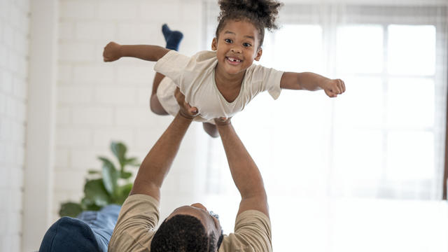 I'm Supergirl!. Happy African cute girl playing with her father on sofa at home. Family happily spend time together. 