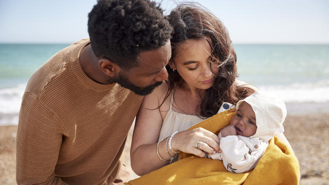 Mother and father, looking adoringly at their newly born baby daughter, on the beach enjoying her first trip to the coast. 