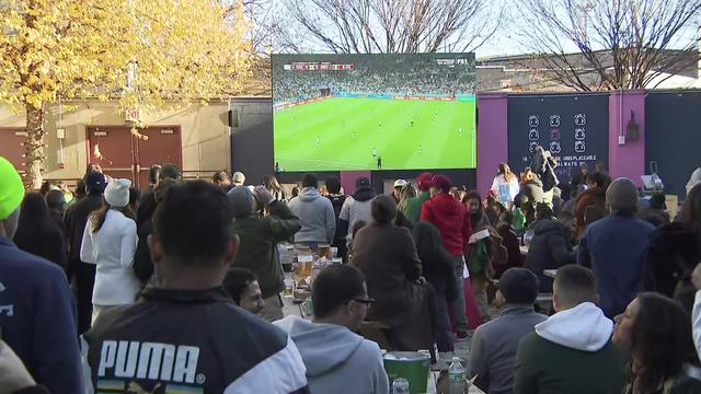 Dozens of people stand and sit in an outdoor patio in front of a jumbotron. 