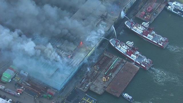 An aerial shot of a large warehouse on the water. Smoke pours from the building as FDNY boats spray water on flames. 