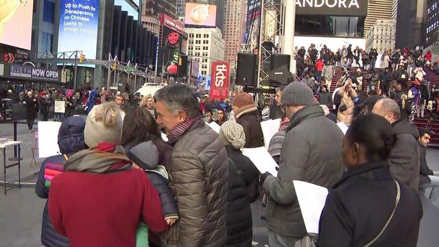 Crowds of people stand in Times Square, looking at sheet music and singing. 